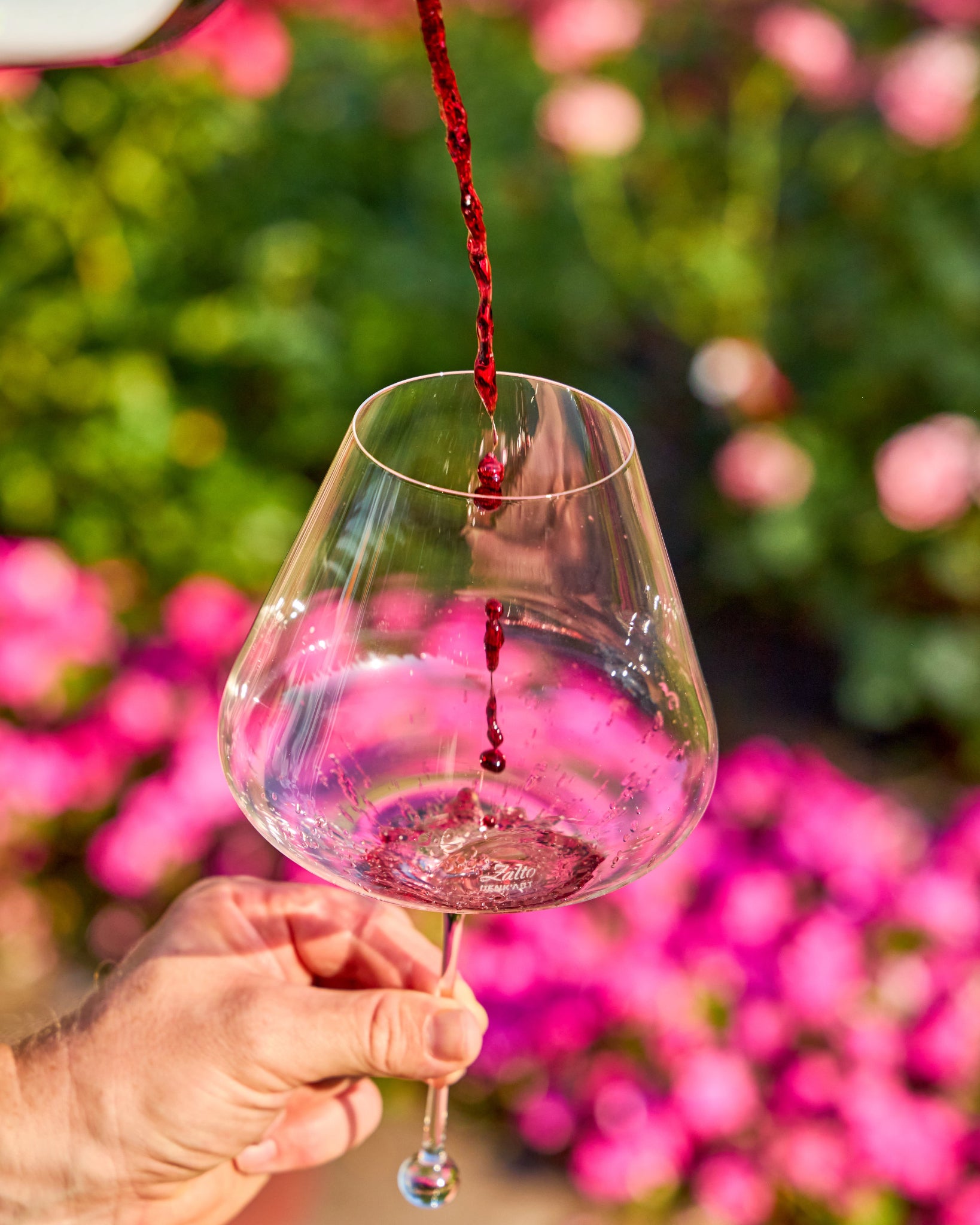 Red wine being poured from a bottle into a clear stemmed Zalto Gravitas Omega glass held by a hand, with a blurred garden background of green foliage and pink flowers.