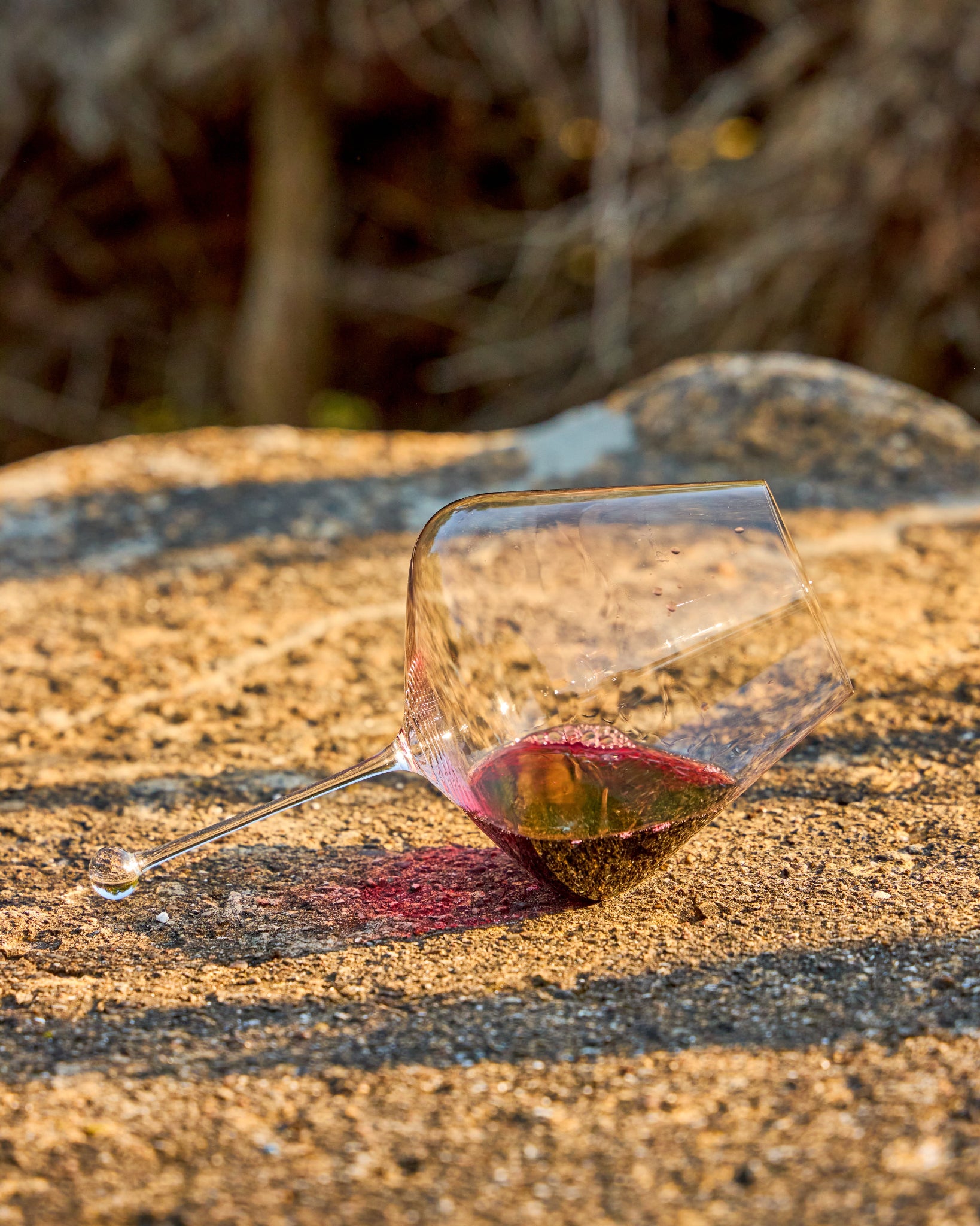 A clear Zalto Gravitas Omega wine glass lying on its side on a sunlit stone surface outdoors, with a small amount of red liquid visible inside and blurred branches in the background.